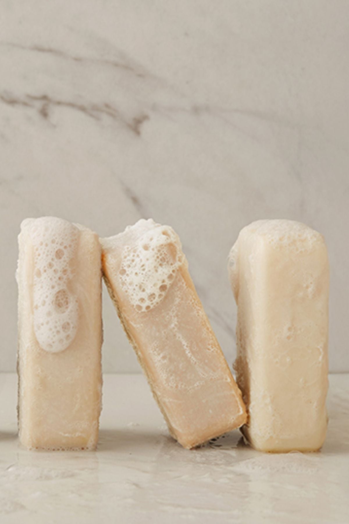 Three pale beige rectangular bars of soap standing on a light marble surface with a matching marble background. One soap is upright on the left with white lather on top, one is leaning in the center with foam on top, and one is upright on the right.