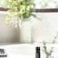 A small black doTERRA essential oil bottle stands on a white surface in a bright, softly blurred bathroom or spa-like setting with plants and a window in the background.