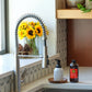 A modern kitchen scene featuring a stainless steel gooseneck faucet with a pull-down sprayer, installed in a white countertop next to a sink. A glass vase of sunflowers, pinecones, a white pump bottle, and a dark amber bottle sit on the counter. doTERRA On Guard Foaming Hand Wash Kit