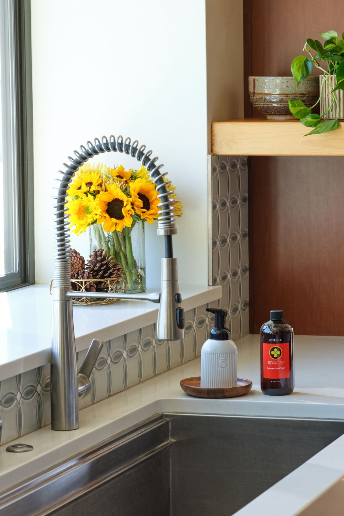 A modern kitchen scene featuring a stainless steel gooseneck faucet with a pull-down sprayer, installed in a white countertop next to a sink. A glass vase of sunflowers, pinecones, a white pump bottle, and a dark amber bottle sit on the counter. doTERRA On Guard Foaming Hand Wash Kit