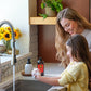 A woman and a young girl wash their hands together at a kitchen sink. The girl wears a yellow knit sweater, and the scene includes soap dispensers, a faucet, flowers, and a plant on a wooden shelf in the background. doTERRA On Guard Foaming Hand Wash Kit