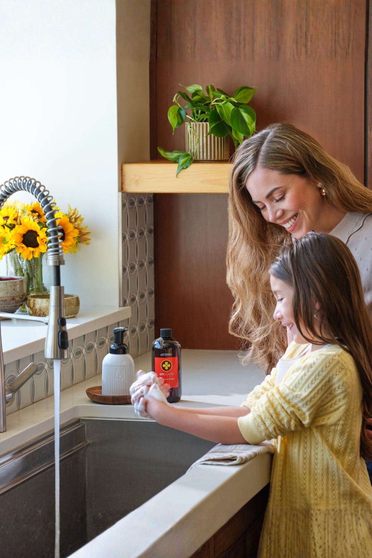 A woman and a young girl wash their hands together at a kitchen sink. The girl wears a yellow knit sweater, and the scene includes soap dispensers, a faucet, flowers, and a plant on a wooden shelf in the background. doTERRA On Guard Foaming Hand Wash Kit