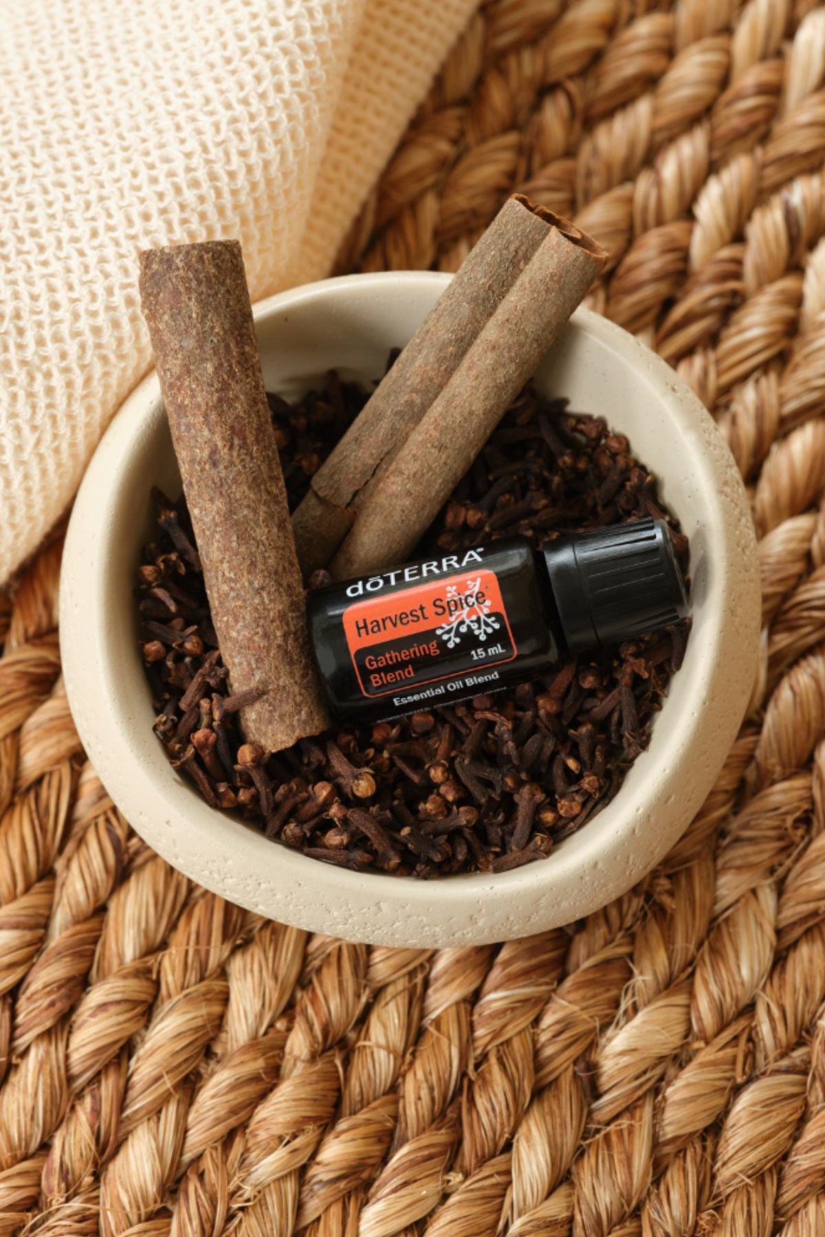 A small black and orange DoTERRA Harvest Spice essential oil bottle rests in a beige bowl filled with cinnamon sticks and cloves, set on a woven straw mat.