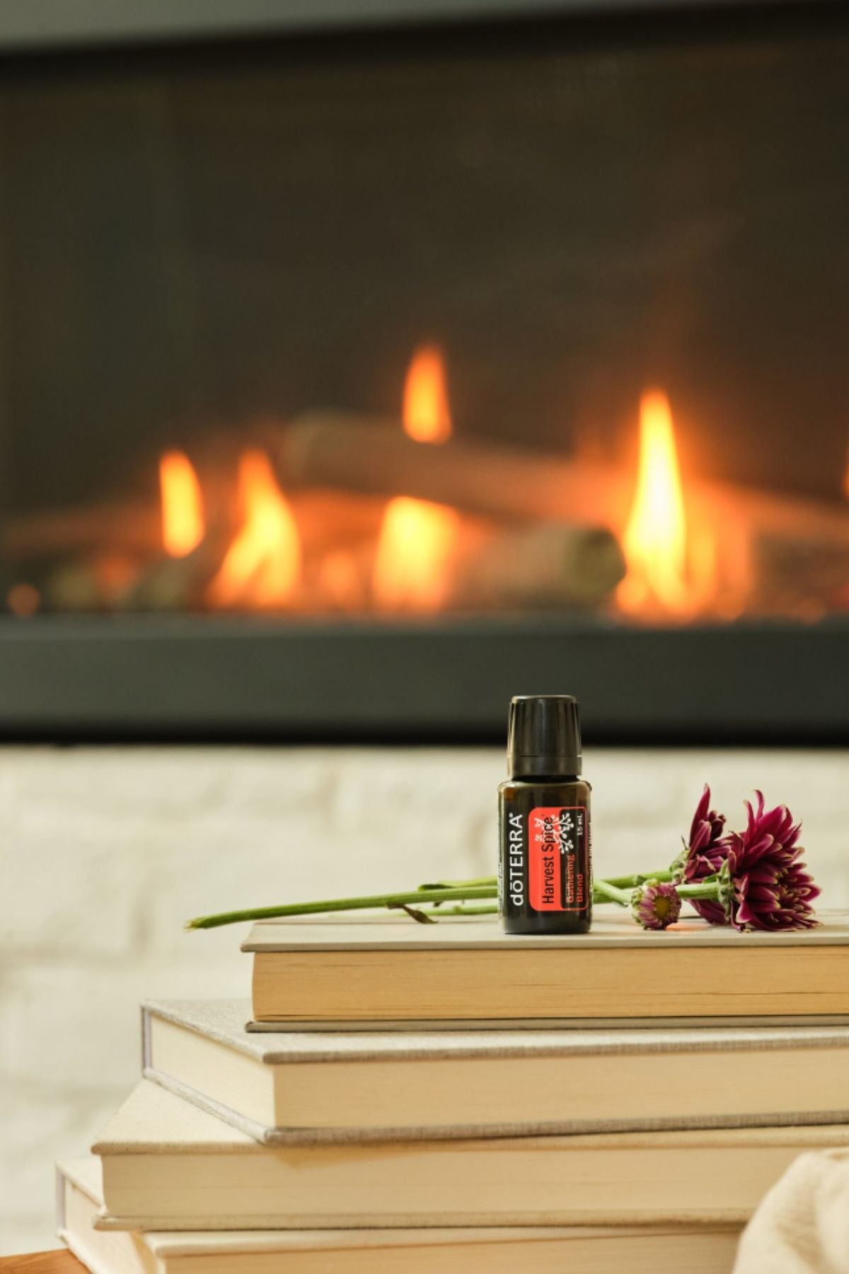 A small bottle of doTERRA cat spray placed on a stack of books in front of a warm fireplace, with a flower resting beside it.