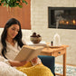 A woman sits in a cozy living room by a fireplace, reading a book wrapped in a chunky mustard yellow knit blanket. A wooden side table beside her holds a white diffuser, a small bowl, and books, with a white brick wall and lit fireplace in the background.