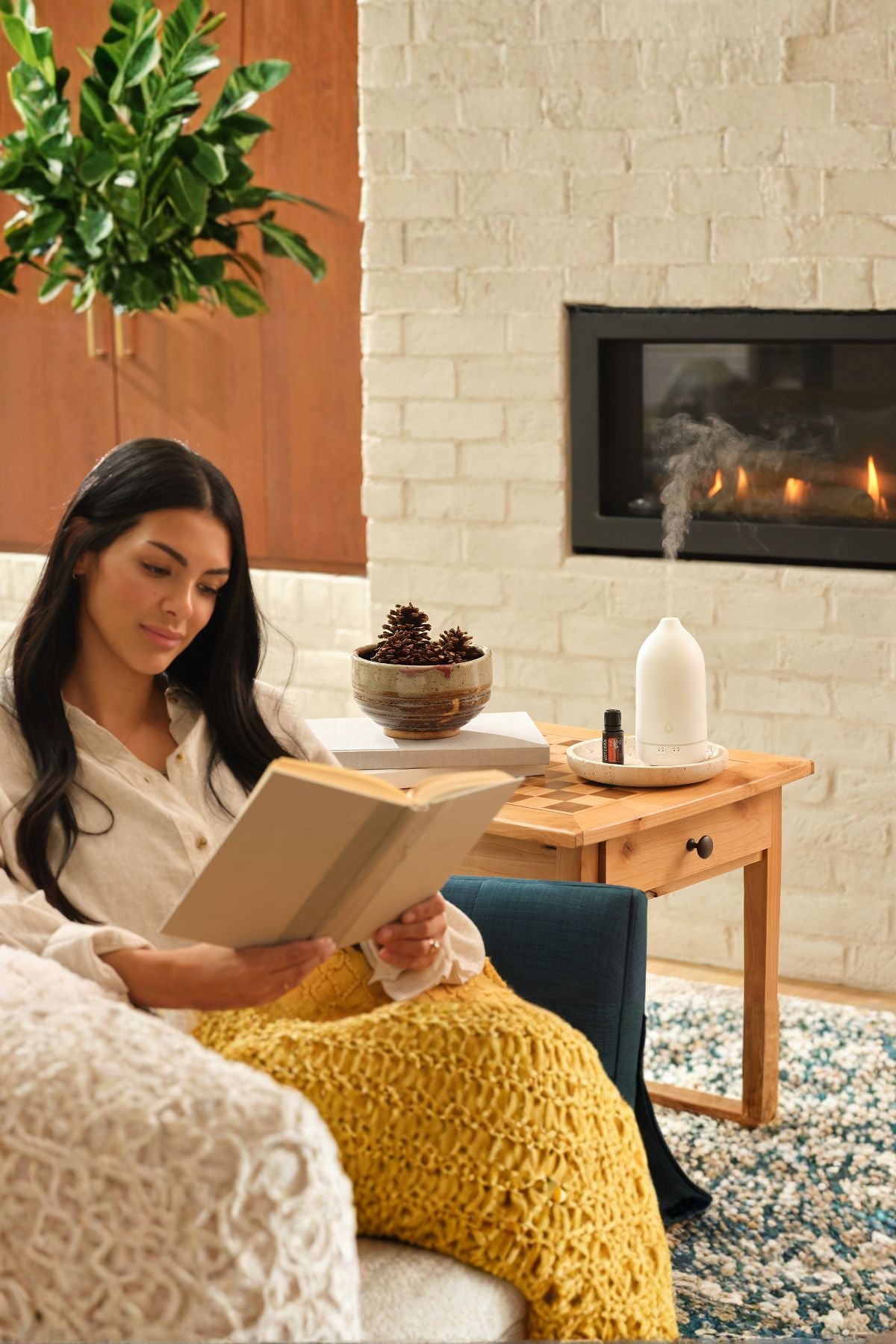 A woman sits in a cozy living room by a fireplace, reading a book wrapped in a chunky mustard yellow knit blanket. A wooden side table beside her holds a white diffuser, a small bowl, and books, with a white brick wall and lit fireplace in the background.
