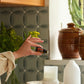 A hand holds a small amber essential oil bottle in a kitchen setting with patterned gray-green tile backsplash, ceramic pots, a white diffuser, and a candle on a woven placemat.