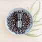 A small black essential oil bottle labeled doTERRA SCULPTOR sits on top of a bowl filled with dark dried botanicals, arranged on a light wood surface with green sprigs beside it. doTERRA Juniper Berry Essential Oil