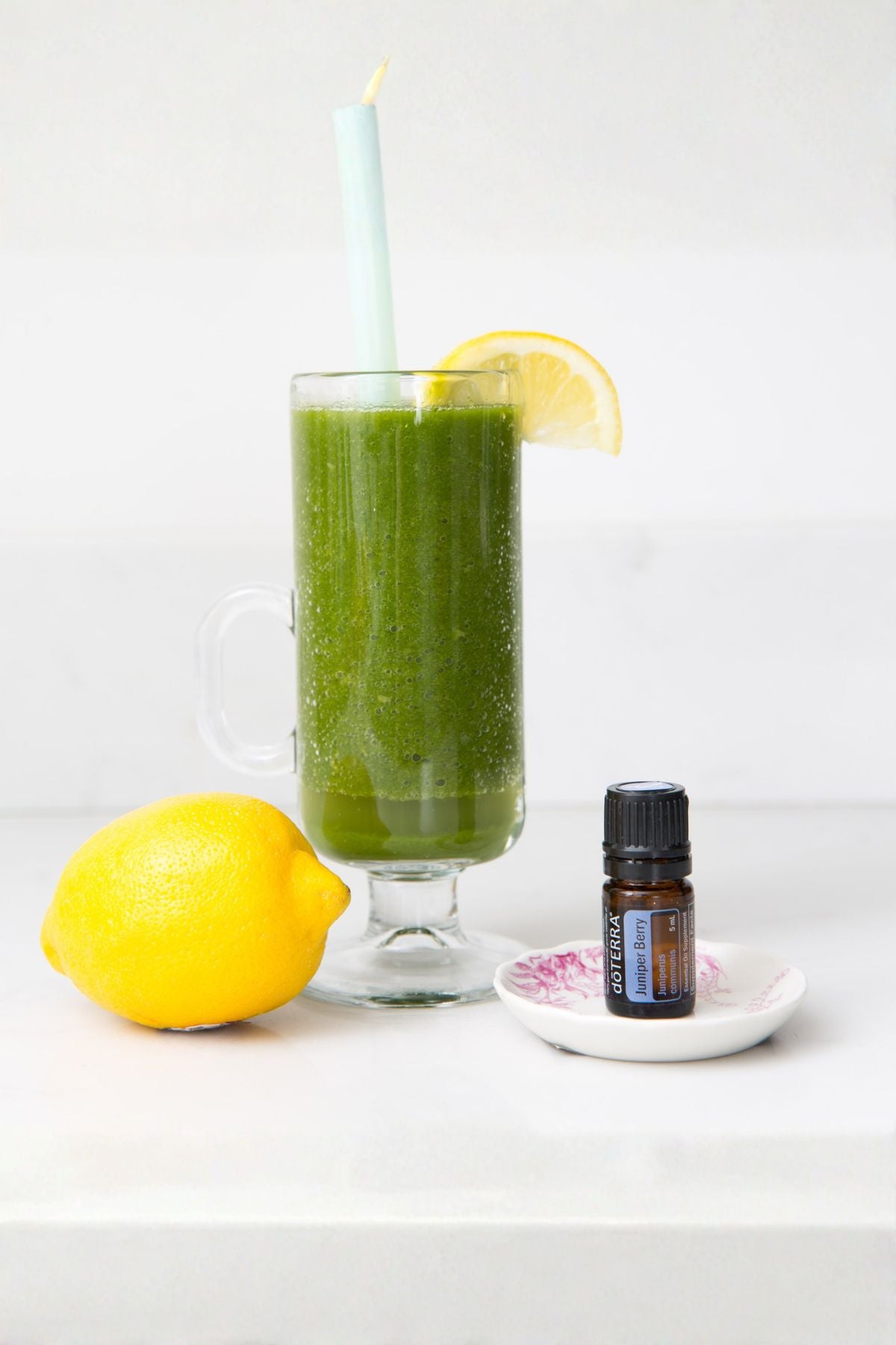 A tall glass mug filled with a bright green juice, set on a white surface with a fresh lemon, a small essential oil bottle, and a candle on a white background. doTERRA Juniper Berry Essential Oil