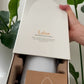 A hand holding the lid of a white Laluz diffuser box, revealing a white diffuser bottle and a brown instruction card inside a brown foam insert, with green plant leaves in the background. dōTERRA LaLuz Diffuser