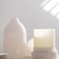 A white diffuser and its matching bottle on a light wooden surface, with soft window light casting diagonal shadows on a white wall. dōTERRA LaLuz Diffuser