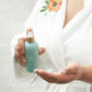 A person in a white robe with a floral print holds a mint green spray bottle with a rose gold sprayer, with their other hand open to receive the mist, against a white tiled wall. dōTERRA Leave-In Conditioner
