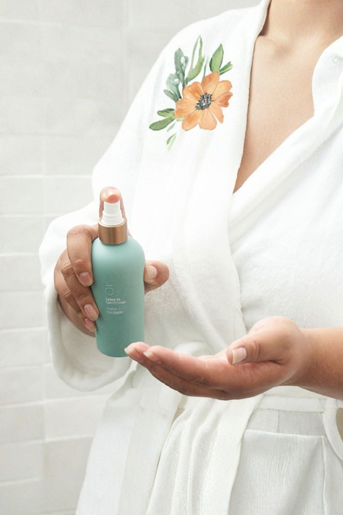 A person in a white robe with a floral print holds a mint green spray bottle with a rose gold sprayer, with their other hand open to receive the mist, against a white tiled wall. dōTERRA Leave-In Conditioner