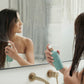 A woman in a bathroom holds a mint green spray bottle in front of a mirror, with her reflection visible. The scene includes a brass shower fixture and soft neutral curtains in the background. dōTERRA Leave-In Conditioner