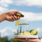 A person's hands are shown adding essential oil from a small amber bottle to a glass of water with lime slices, held on a tray outdoors against a blurred blue sky and water background. doTERRA Lime Essential Oil