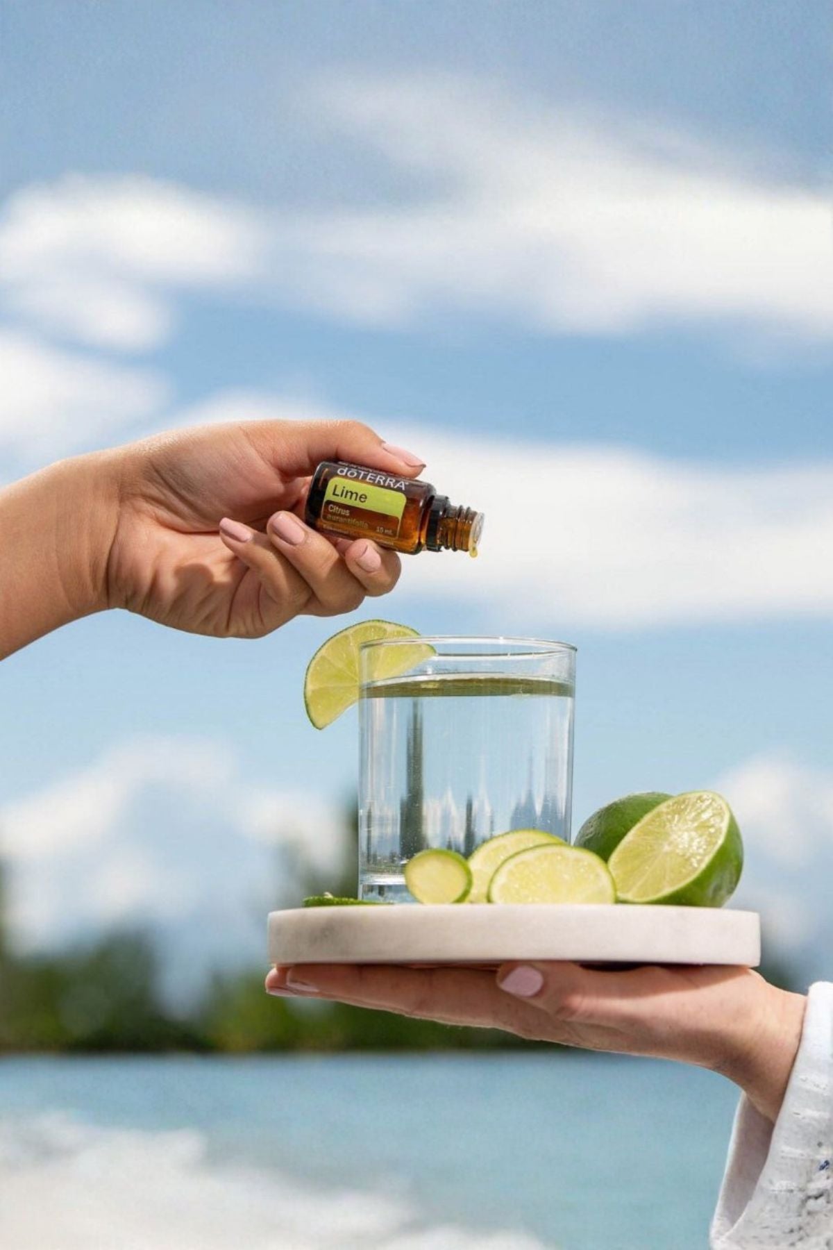 A person's hands are shown adding essential oil from a small amber bottle to a glass of water with lime slices, held on a tray outdoors against a blurred blue sky and water background. doTERRA Lime Essential Oil