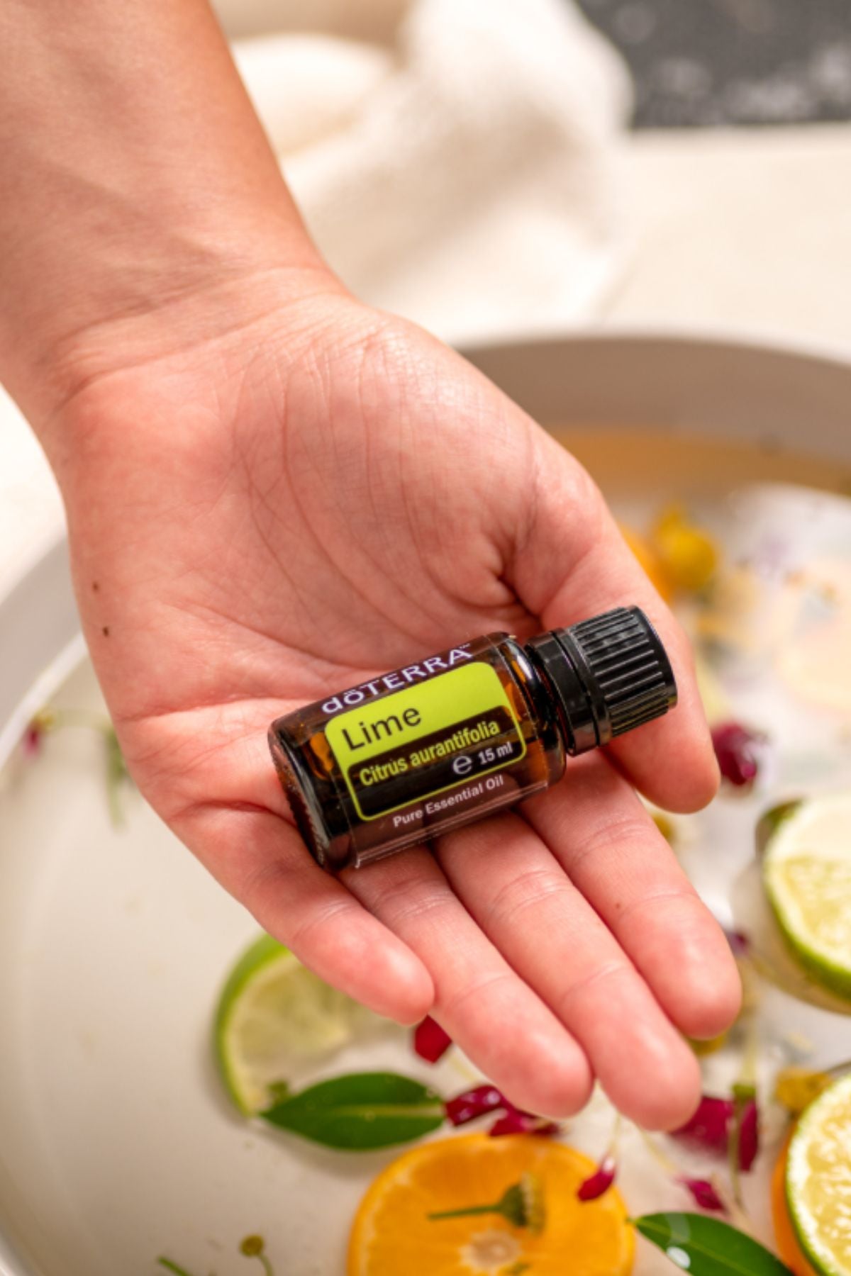 A hand holding a small bottle of doTERRA Lime Cellar Botanicals essential oil over a bowl of citrus slices and herbs.
