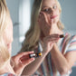 A woman in a striped shirt applies doTERRA Melissa oil to her face while looking into a mirror, holding a small amber bottle in the foreground. 