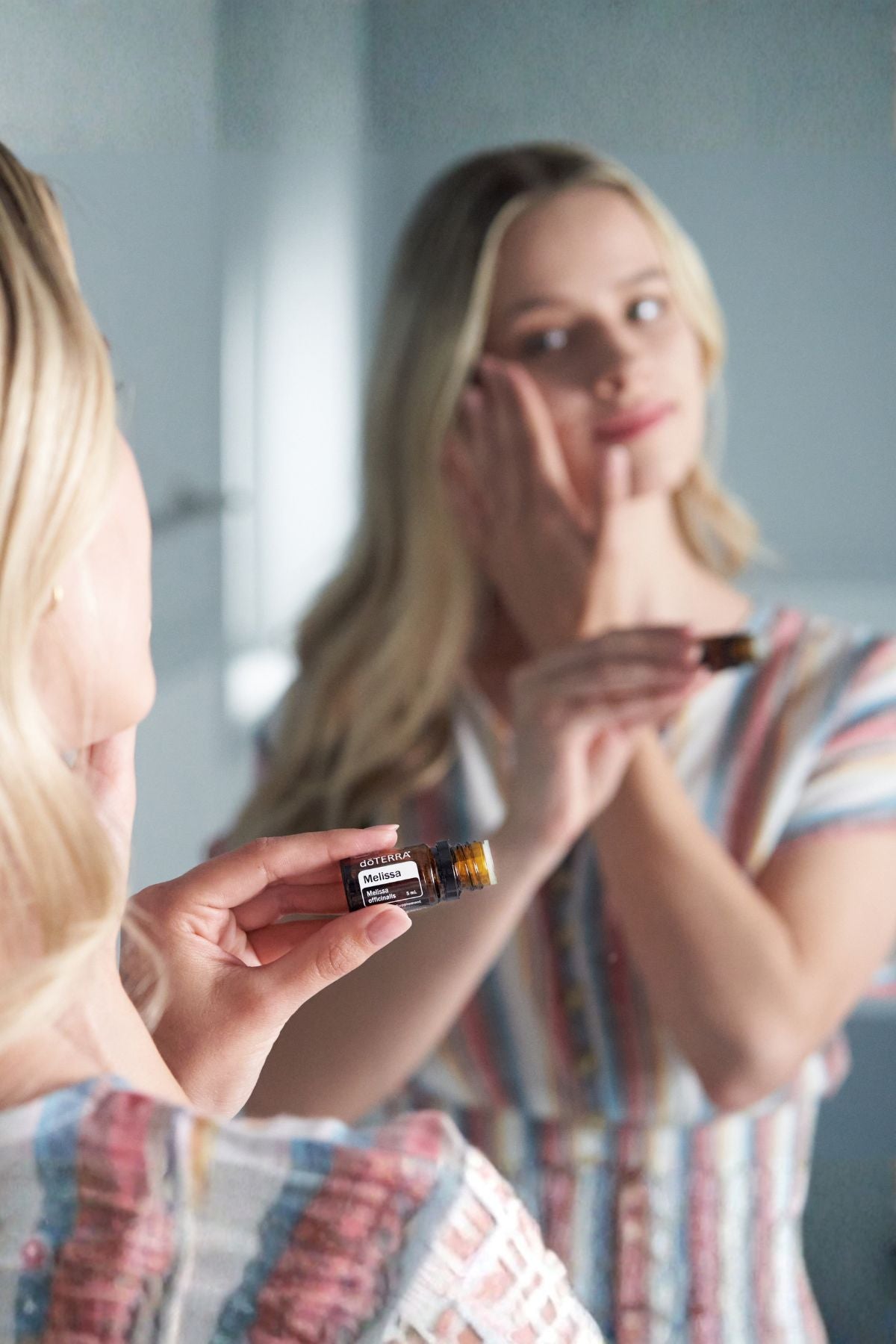 A woman in a striped shirt applies doTERRA Melissa oil to her face while looking into a mirror, holding a small amber bottle in the foreground. 