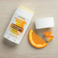 A doTERRA Natural Sensitive Deodorant stick with its cap removed, placed on a light wood surface next to an orange slice and orange peel. The product packaging is white with orange and yellow text, and the deodorant stick itself is visible at the top.