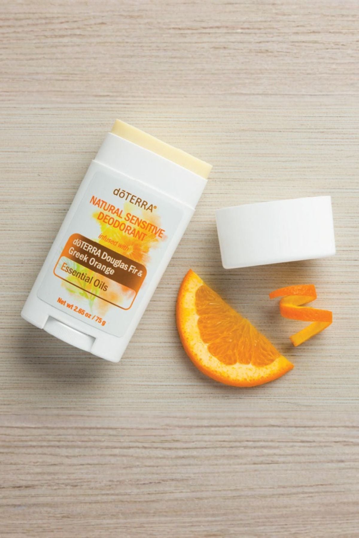 A doTERRA Natural Sensitive Deodorant stick with its cap removed, placed on a light wood surface next to an orange slice and orange peel. The product packaging is white with orange and yellow text, and the deodorant stick itself is visible at the top.