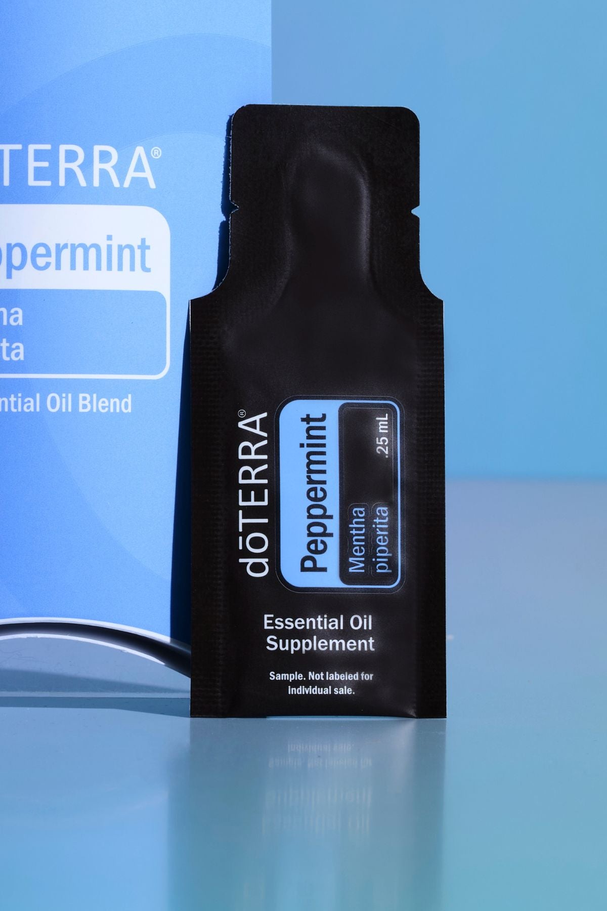 A single black dOTERRA Peppermint Essential Oil Supplement sachet standing upright on a glossy blue surface, with a blue branded box partially visible behind it. doTERRA Peppermint Essential Oil Sample (.25 mL)
