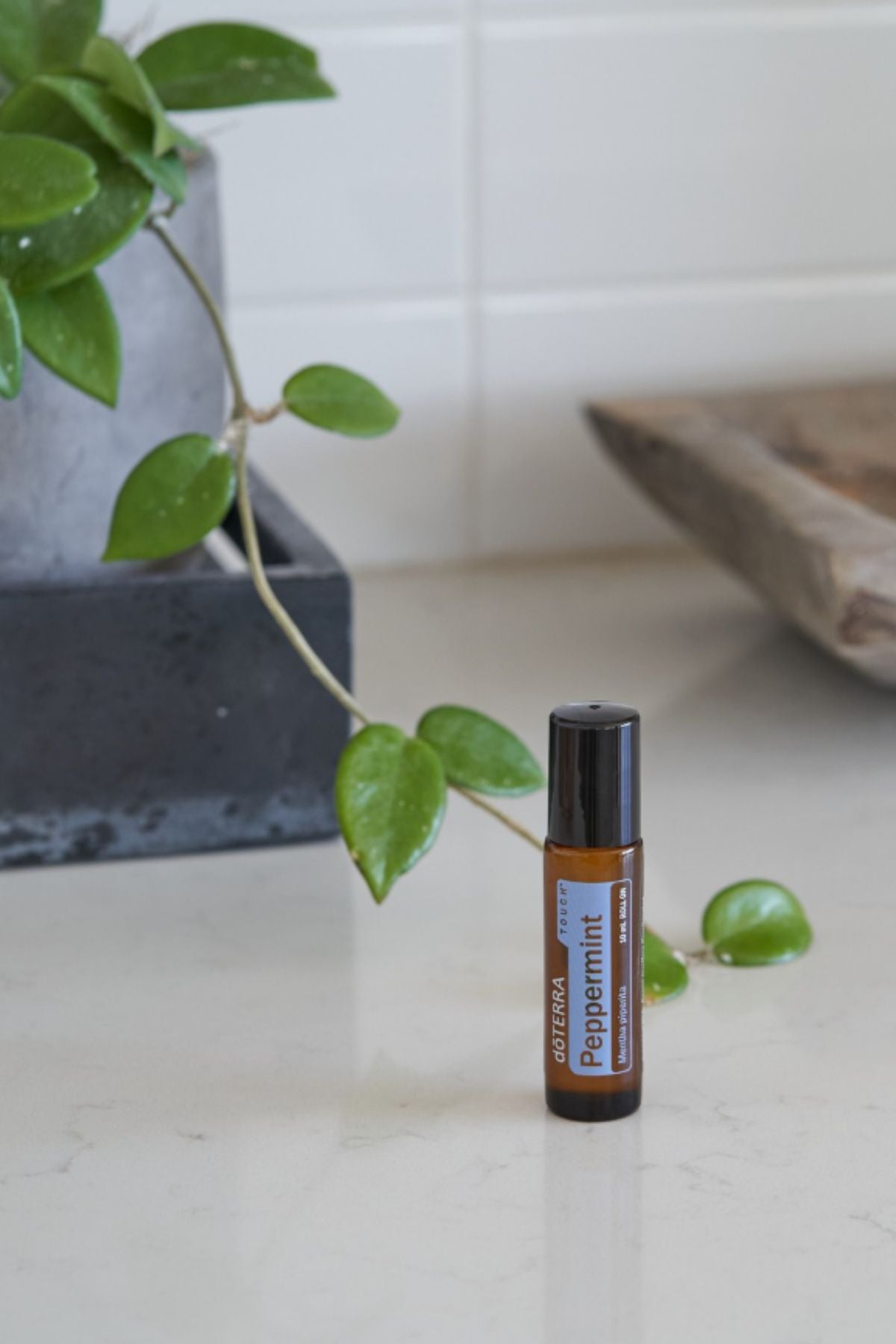 A small amber roll-on bottle labeled doTERRA Peppermint sits on a light countertop, with a trailing green plant and a neutral interior background visible.