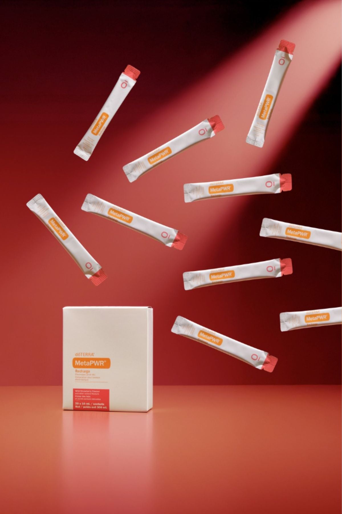 A white rectangular box labeled MegaPWT stands on a red surface, with multiple white single-serve packets floating around it against a red studio background. doTERRA MetaPWR Recharge Wild Strawberry Electrolytes
