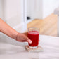 A hand holding a clear glass filled with bright red juice, resting on a square concrete coaster on a white marble countertop in a bright kitchen setting. doTERRA MetaPWR Recharge Wild Strawberry Electrolytes