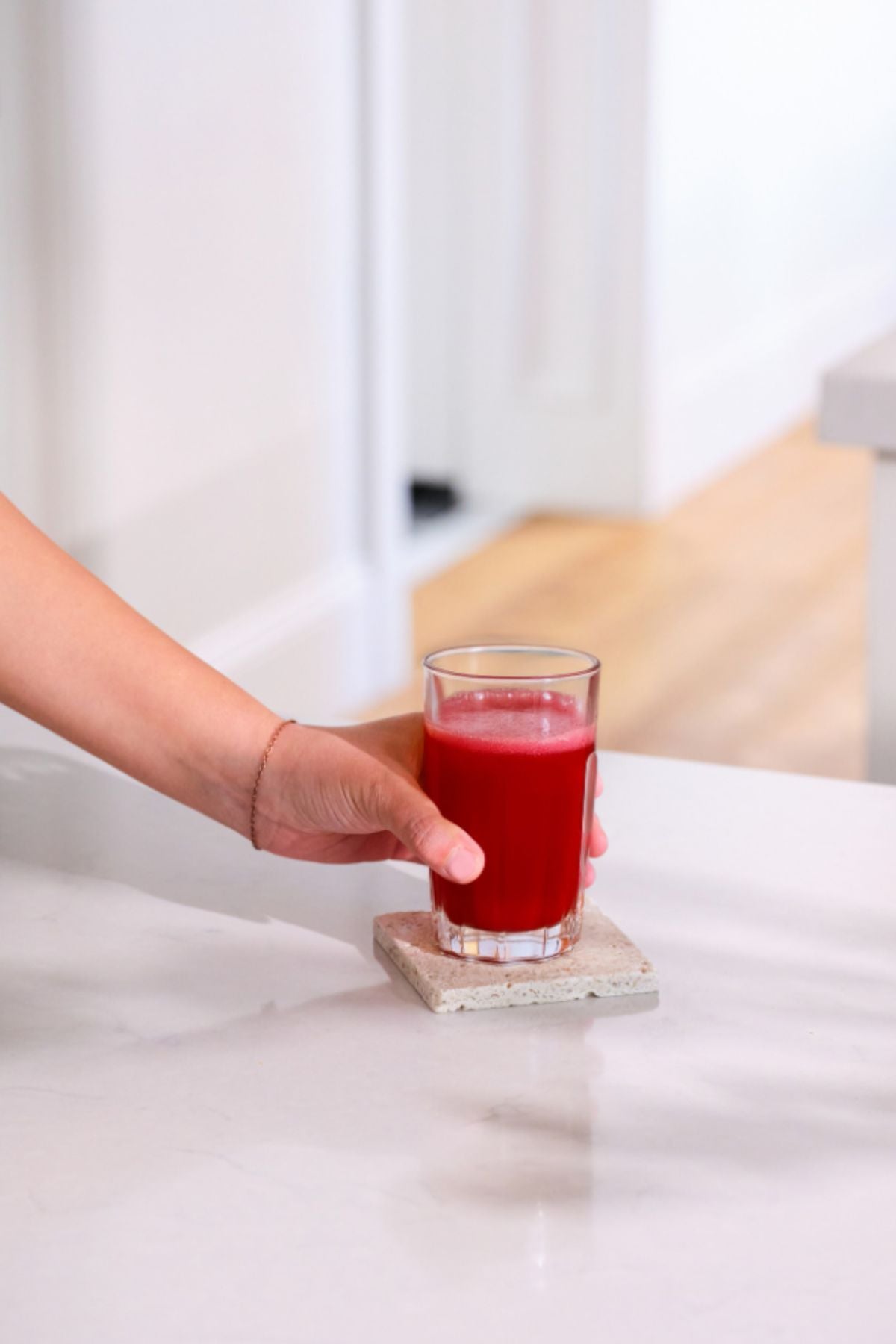 A hand holding a clear glass filled with bright red juice, resting on a square concrete coaster on a white marble countertop in a bright kitchen setting. doTERRA MetaPWR Recharge Wild Strawberry Electrolytes