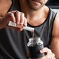 A man in a gray tank top mixes a red liquid into a clear shaker bottle with a black sleeve, holding a white and orange MicroPlenty packet above the bottle. doTERRA MetaPWR Recharge Wild Strawberry Electrolytes