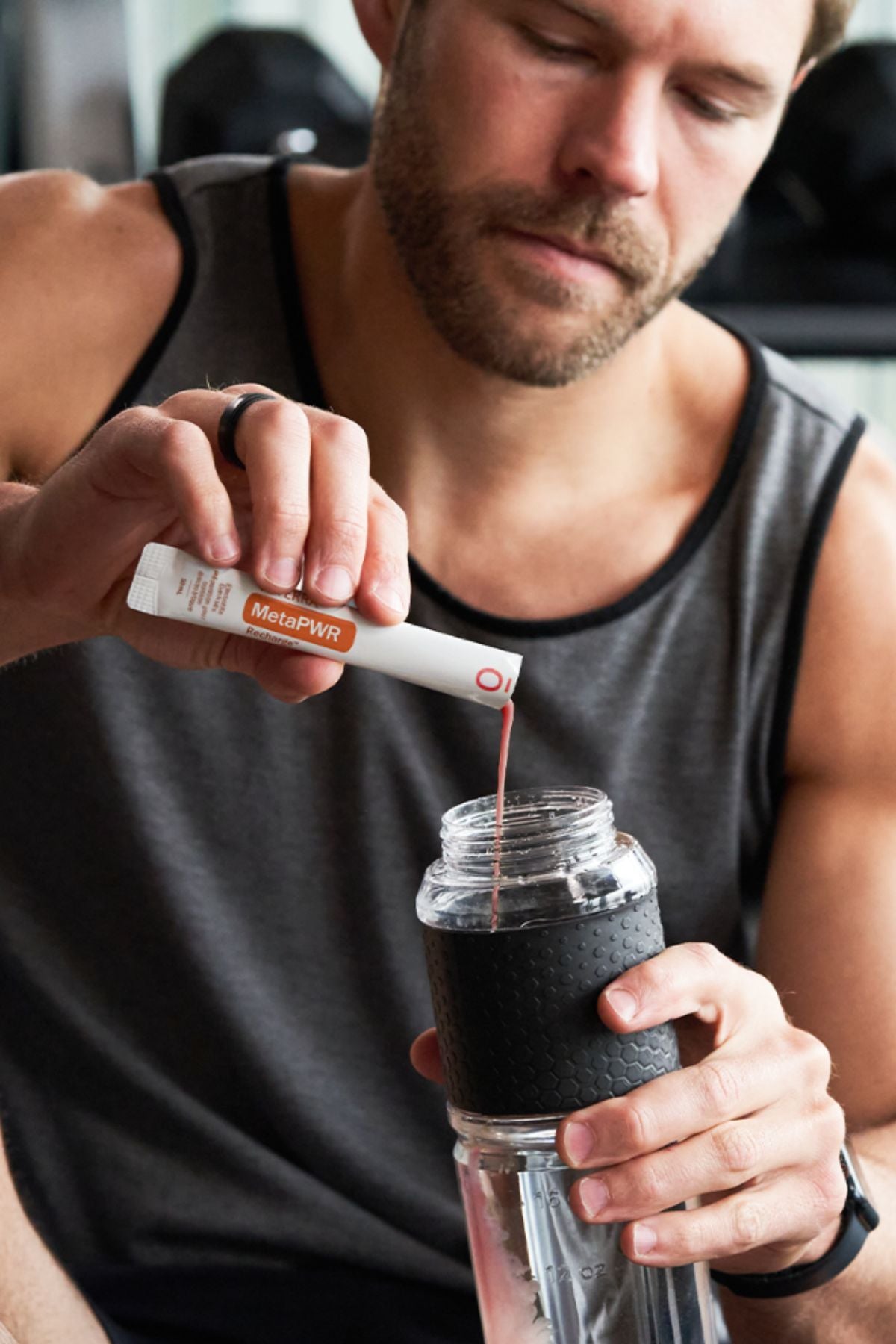 A man in a gray tank top mixes a red liquid into a clear shaker bottle with a black sleeve, holding a white and orange MicroPlenty packet above the bottle. doTERRA MetaPWR Recharge Wild Strawberry Electrolytes
