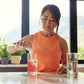 A woman in an orange sleeveless top stands at a kitchen counter pouring a pink drink into a glass, with two clear glasses and a bright window view of mountains in the background. doTERRA MetaPWR Recharge Wild Strawberry Electrolytes