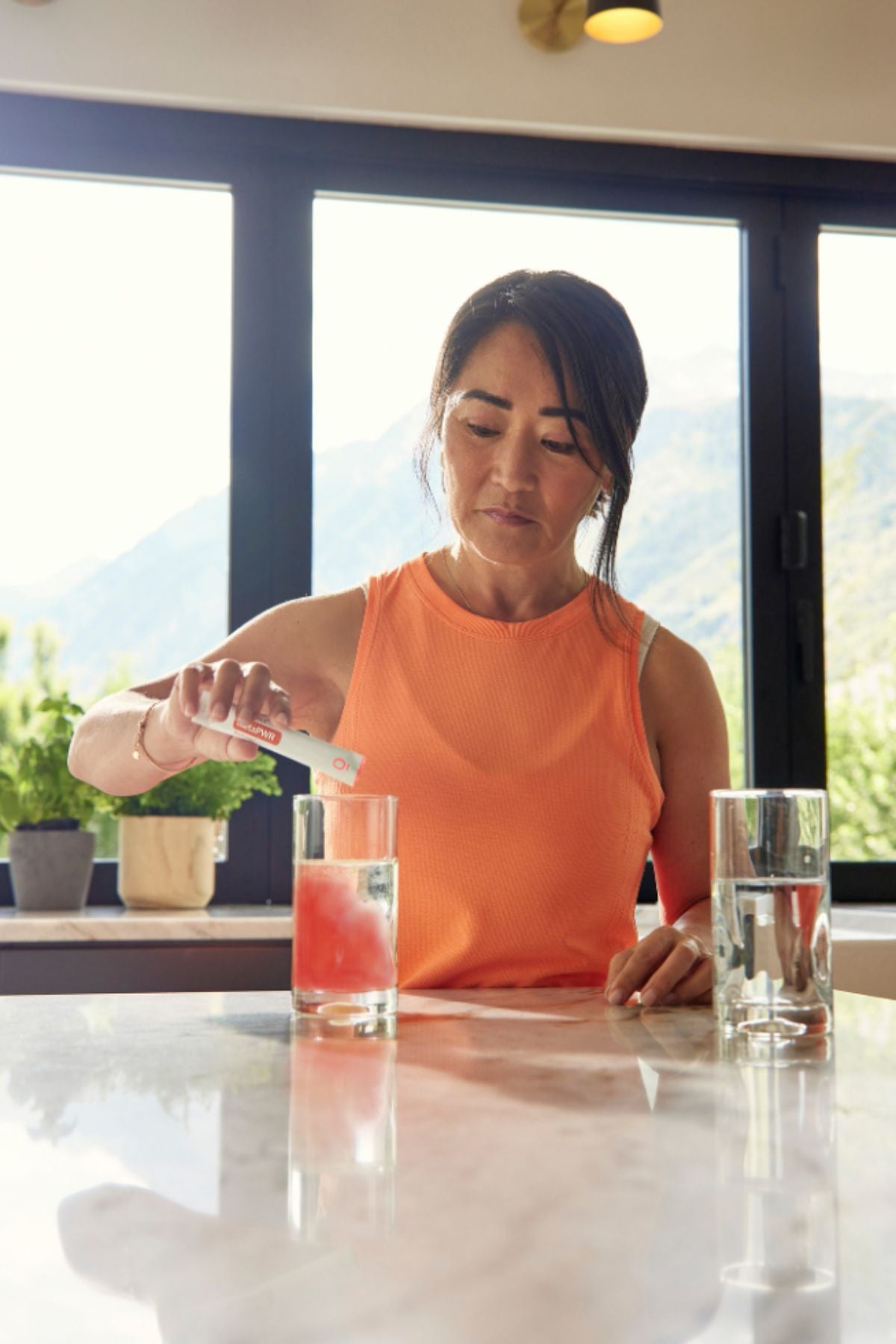 A woman in an orange sleeveless top stands at a kitchen counter pouring a pink drink into a glass, with two clear glasses and a bright window view of mountains in the background. doTERRA MetaPWR Recharge Wild Strawberry Electrolytes