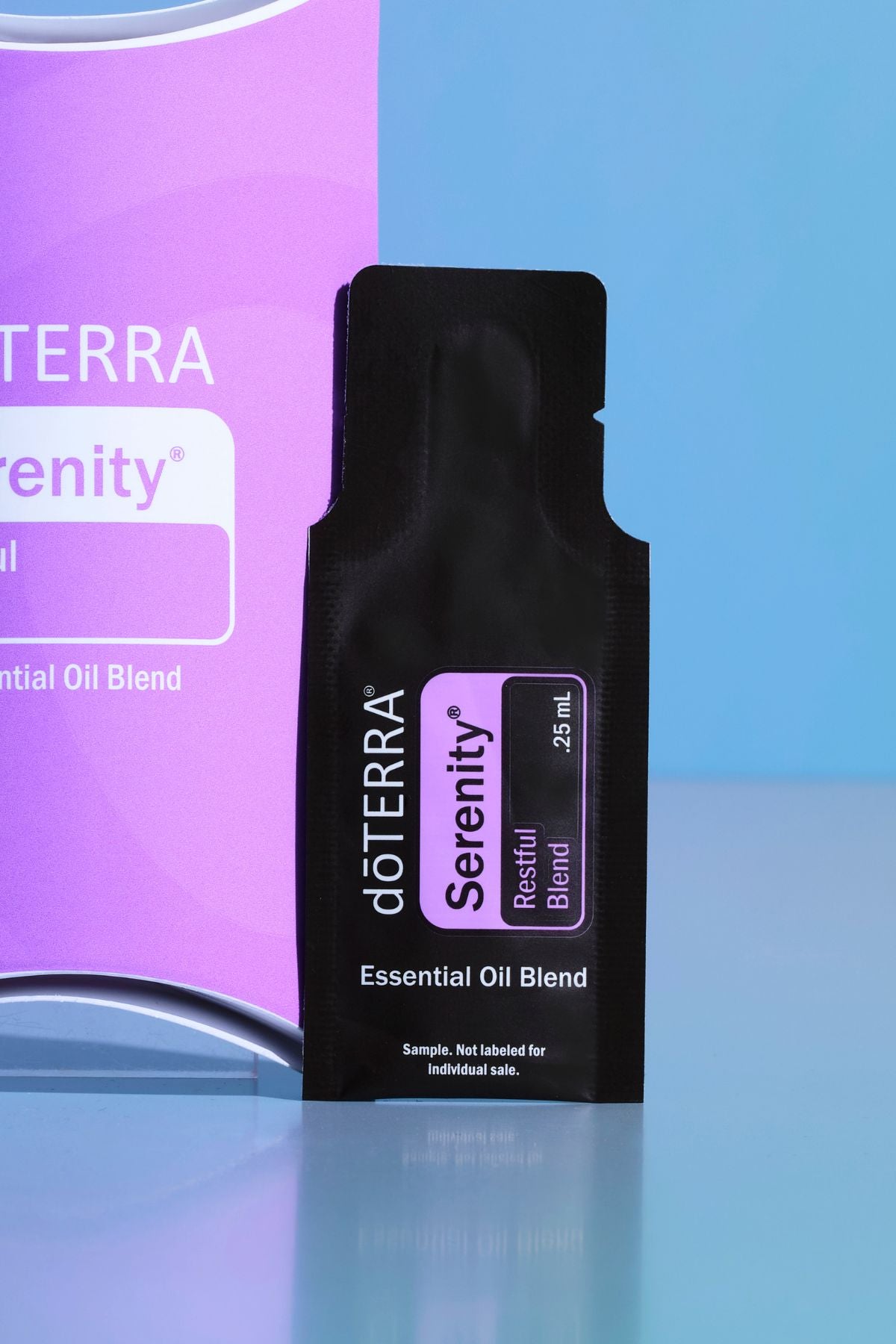 A black doTERRA Serenity Restful Blend essential oil blend sample sachet standing upright in front of a purple and blue branded box. doTERRA Serenity Essential Oil Sample (.25 mL)