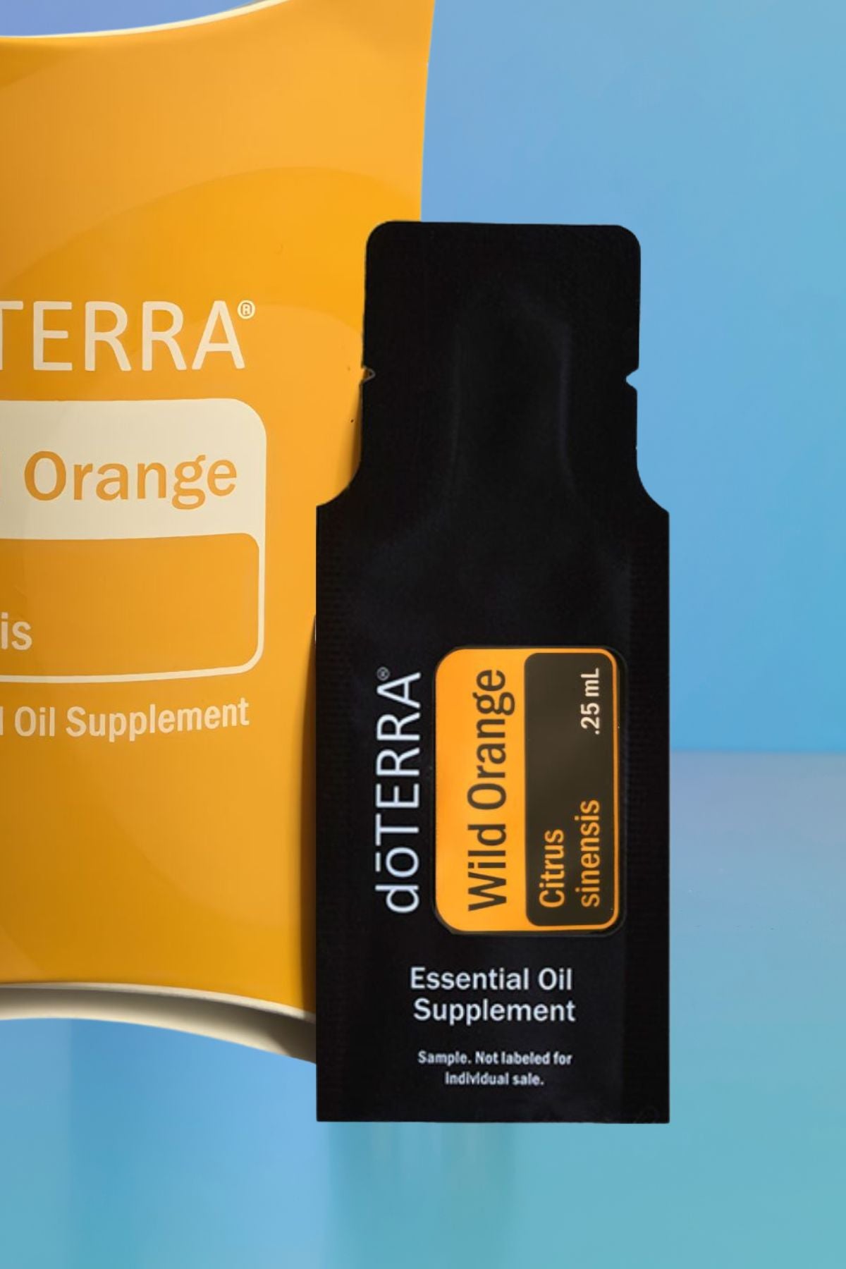 A black doTERRA Wild Orange citrus slice essential oil supplement sachet in front of a yellow-orange doTERRA box on a blue gradient background. doTERRA Wild Orange Essential Oil Sample (.25 mL)
