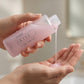 A person's hands holding a clear plastic bottle of Asotebra IKISE Lotion, Shower & Body Wash, with pink gel being poured from the bottle into the other hand.