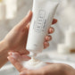 A person's hands holding a white tube of Juvéderma Uplift Cream 2 Face Wash, with the foam being squeezed into another hand against a soft, blurred bathroom background.