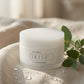 A jar of doTERRA IKISE 1 Cleansing Balm sits on a beige textured fabric surface with water droplets, next to green eucalyptus leaves, against a soft, warm neutral background.