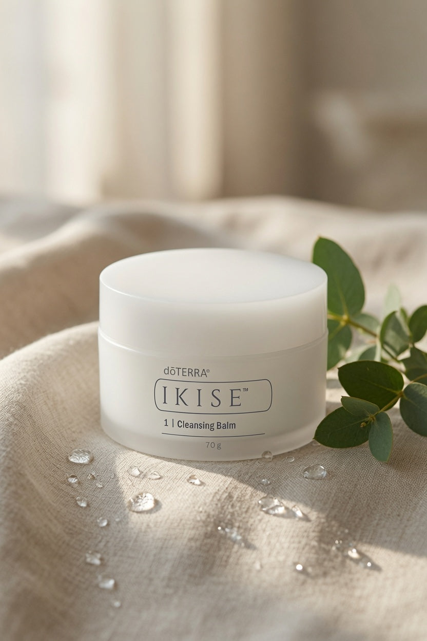 A jar of doTERRA IKISE 1 Cleansing Balm sits on a beige textured fabric surface with water droplets, next to green eucalyptus leaves, against a soft, warm neutral background.